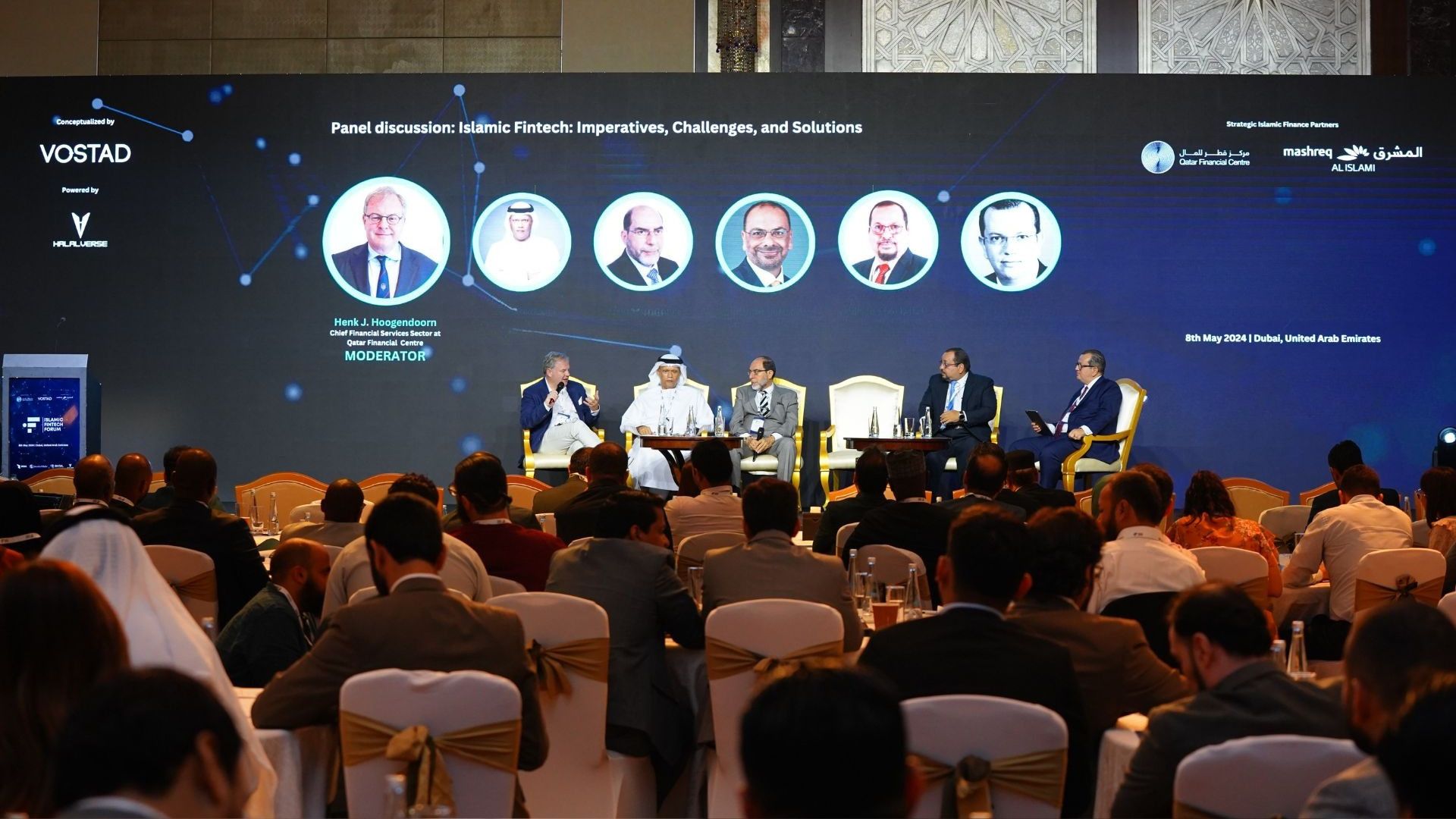 A panel session under way on stage before a seated audience at a Vostad forum